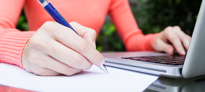 A woman writing on a a paper with the laptop on the side