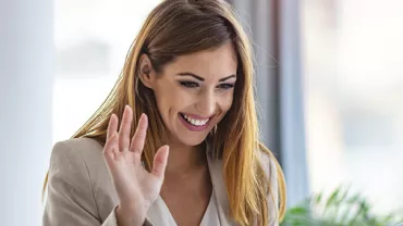 Woman smiling and waving while looking at papers during a video call.