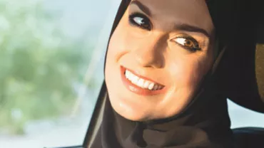 Smiling person wearing a hijab in a car seat, sunlight on face.