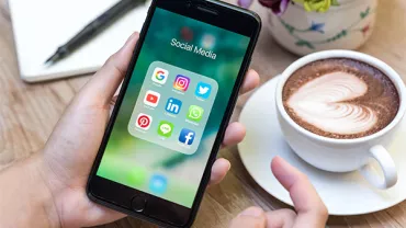 Person holding a smartphone displaying social media apps next to a latte with heart art.