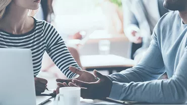 Two people having a discussion at a table with a laptop and coffee cup.