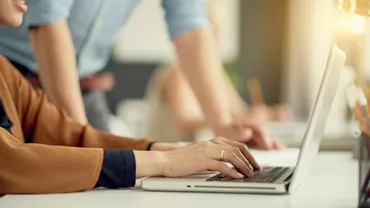 Person typing on a laptop at a desk with another person standing nearby.