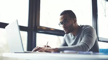 Man wearing glasses working on a laptop near large windows.