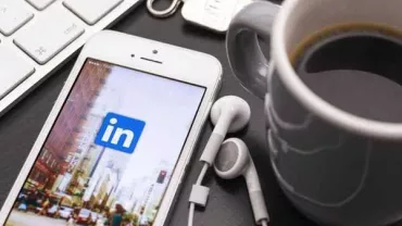 Smartphone with LinkedIn app on screen, beside earbuds, keyboard, and coffee mug.