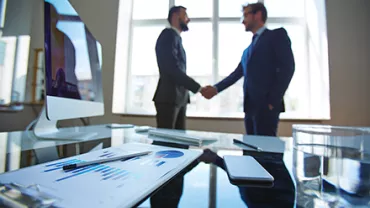 Two businessmen shaking hands in an office with a desktop and charts visible.