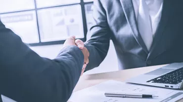 Two businesspeople shaking hands at a desk with documents and a laptop.