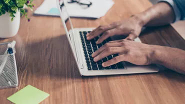 Person typing on a laptop at a wooden desk with papers and a plant.