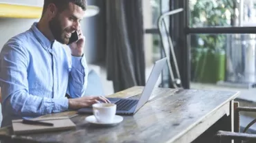 Man talking on phone while working on laptop at a wooden table.