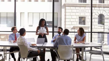 A woman stands and speaks to a group in an office meeting room.