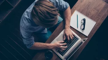 Top view of person typing on laptop at wooden desk with notebook, glasses, and pencil