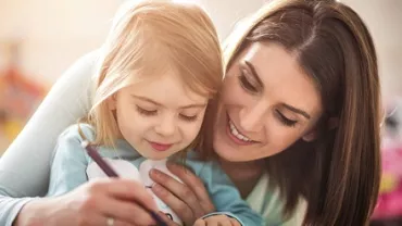 Smiling woman helps young girl draw with a pen.