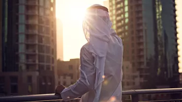 A person in traditional attire stands on a balcony overlooking a cityscape at sunrise.