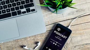 Smartphone on a wooden table playing a podcast beside a laptop and earphones.