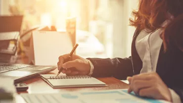 Person writing notes at a desk with sunlight in the background.