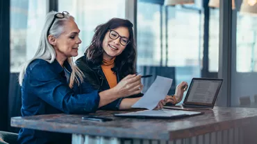 Two women discussing documents at a table with a laptop.