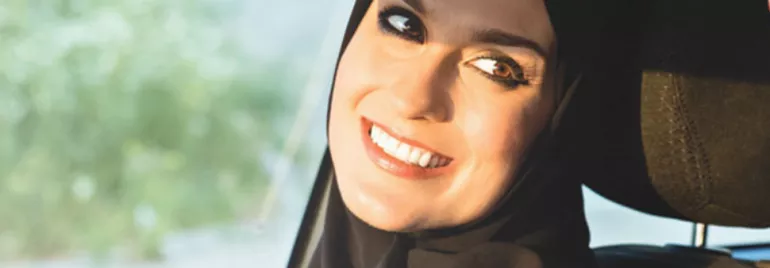 Smiling person wearing a hijab in a car seat, sunlight on face.